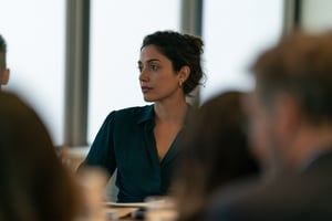 A woman in a teal blouse sits at a meeting table, looking away with a thoughtful, reserved expression while colleagues talk around her.
