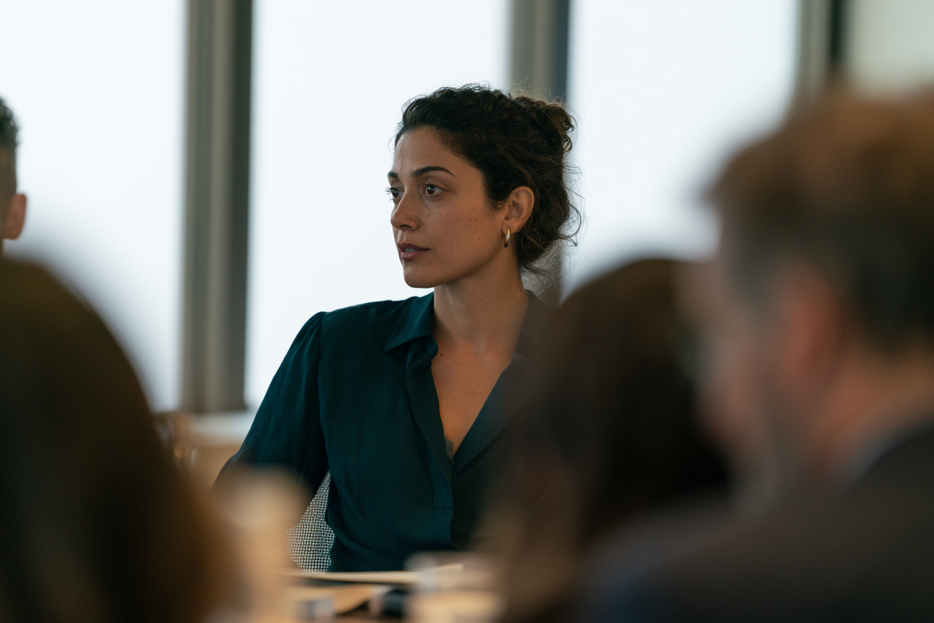A woman in a teal blouse sits at a meeting table, looking away with a thoughtful, reserved expression while colleagues talk around her.
